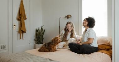 Two women sitting on a bed with a dog. They are having a conversation. 